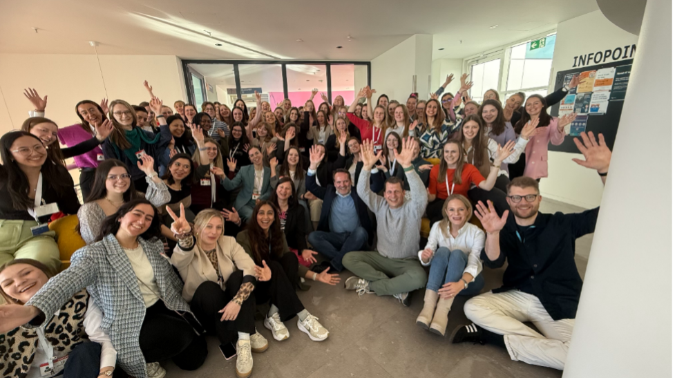 Das Bild zeigt eine große Gruppe von Menschen, die zusammen in einem Innenraum sitzen und stehen. Sie lächeln, winken und wirken fröhlich.