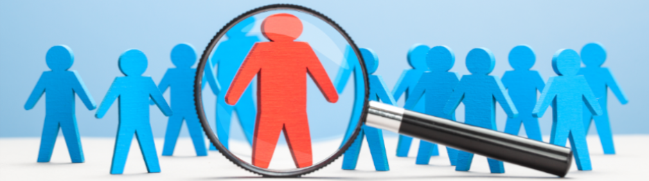 A magnifying glass is focused on the single red figure in a group of otherwise blue paper figures standing on a white surface.
