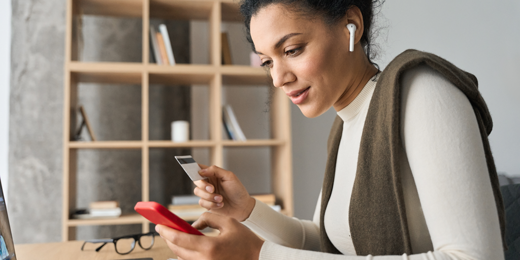 Young adult African American female consumer holding credit card and smartphone sitting at desk at home doing online banking transaction