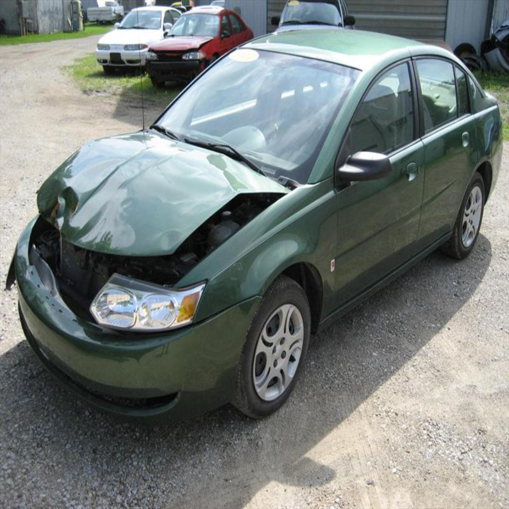 Green sedan with visible front collision damage displayed in automotive repair lot, showing extent of hood and bumper impact