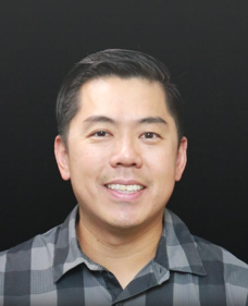 Professional headshot of a smiling Asian man with short dark hair wearing a gray and black checkered collared shirt against a dark background
