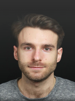 Professional headshot of a young man with brown hair and facial stubble wearing a gray t-shirt against a dark background