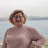 Portrait of a smiling person wearing round glasses and a pink knitted sweater, posing outdoors by a waterfront with a scenic lake background.