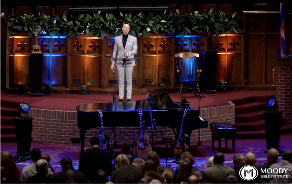 Screenshot from Moody Bible Institute of a television screen depicting a man on stage leading service for a large audience.