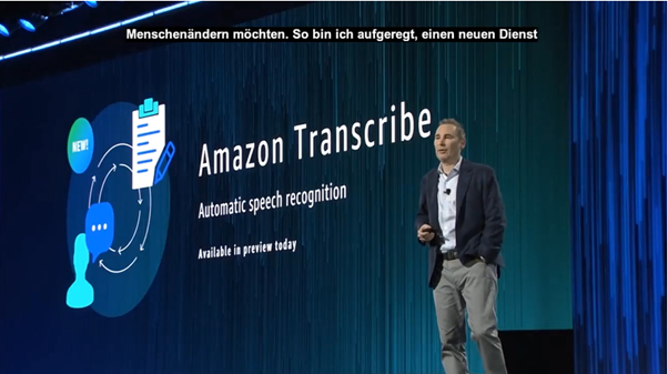 Example of captions showing on the top of video. The image is of a speaker on a stage giving a presentation in their native language of English. Copy is displayed on a screen behind them stating: Amazon Transcribe, Automatic speech recognition, Available in preview today. Across the top of the image is closed captioning in German which reads as: Menschenandern mochten. So bin ich aufgeregt, einen nauen Dienst.